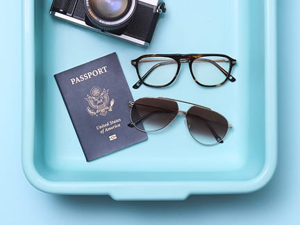 A security screening tray containing a pair of metal aviator sunglasses and a pair of tortoiseshell eyeglasses, a passport, a Zenni glasses case, and a vintage film camera.