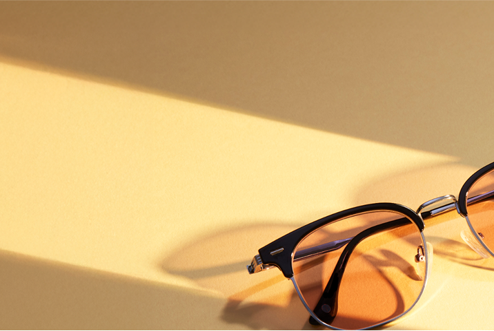 A vintage-inspired pair of browline sunglasses featuring warm amber-tinted lenses. The glossy black upper rim contrasts beautifully with the sleek metal frame, casting soft shadows on a golden background.