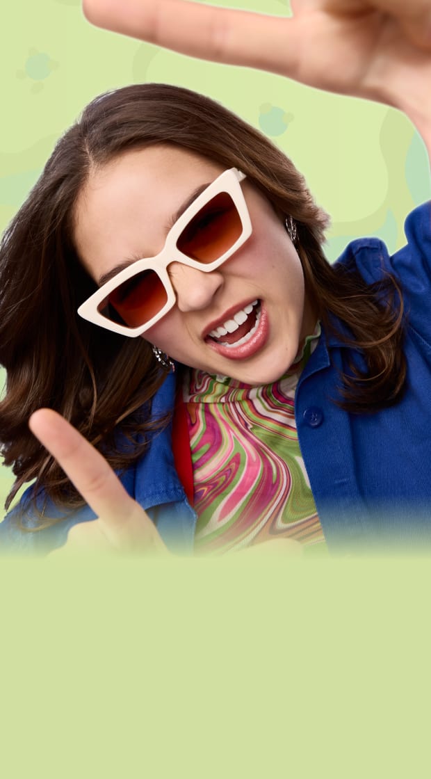 A young woman with long, wavy brown hair wears bold white cat-eye sunglasses with dark lenses. She is dressed in a groovy, multicolored swirl-patterned top layered under a vibrant blue jacket, against a pastel green with abstract organic shapes background.