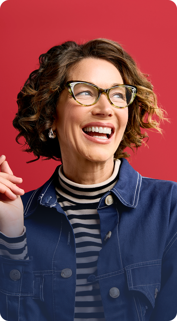 Smiling woman with short curly hair wearing bold cat-eye glasses, a striped turtleneck, and a denim jacket, set against a red background.