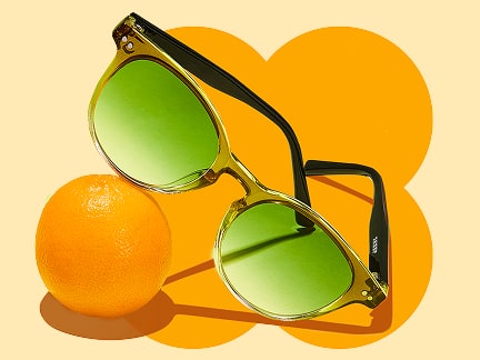 Green round sunglasses with green lenses on an orange background, resting on a small orange.
