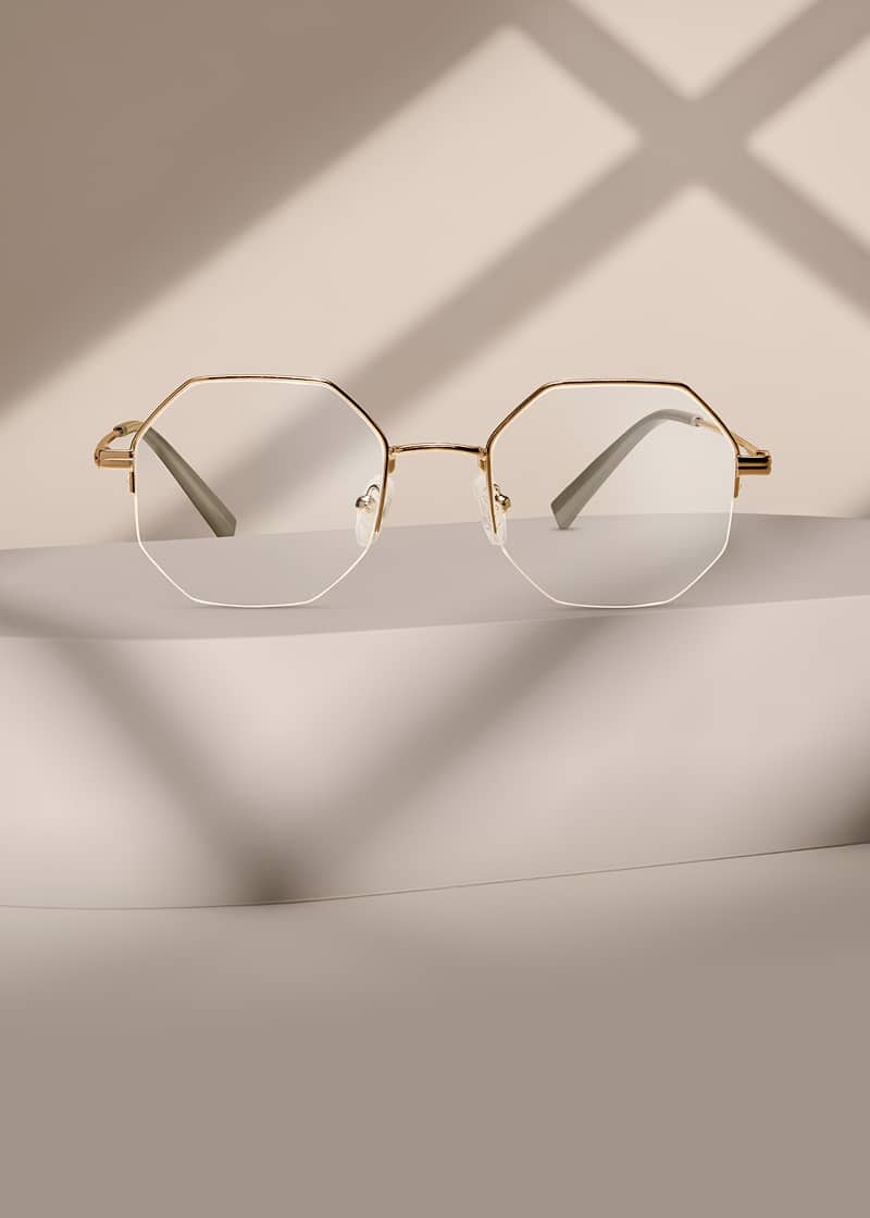 Gold geometric half-rim metal Zenni glasses with clear lenses on a beige pedestal.