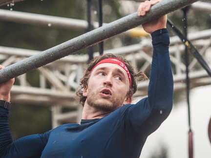 A man wearing a red headband and a navy long-sleeve shirt hangs from a metal bar during an outdoor obstacle course, with rain droplets falling and scaffolding in the background.