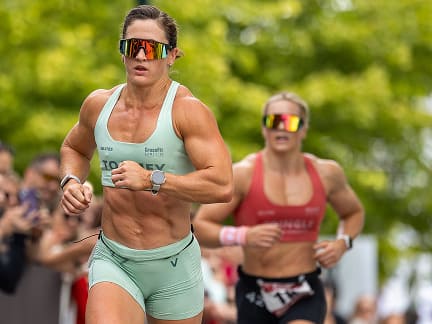 Two athletes wearing reflective sunglasses run outdoors, one in green in the foreground and one in red behind, with blurred trees and spectators in the background.