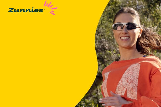 A smiling woman runs outdoors wearing wraparound sunglasses and an orange sweatshirt. 