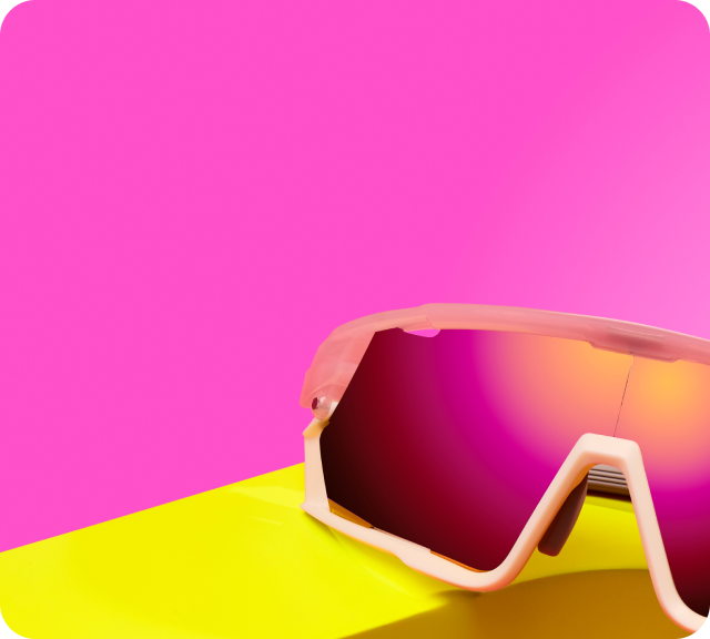 A pair of sporty wraparound sunglasses with a pink frame and vibrant mirrored lenses sits on a bright yellow surface against a bold pink background with rounded corners.