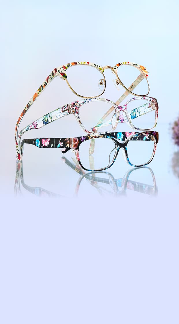 A stack of three floral-patterned eyeglasses is displayed against a soft blue background, with their reflections on a glossy surface and blurred flowers in the background.