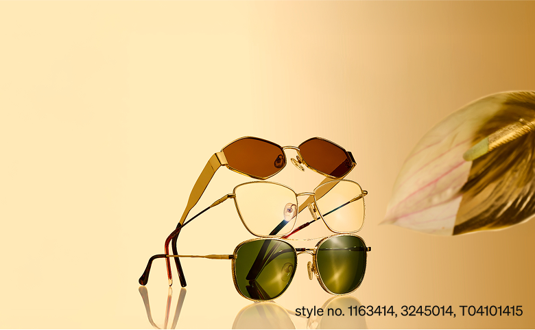 Stack of three gold-framed sunglasses with different lens tints—brown, clear, and green—on a reflective surface against a warm golden background.