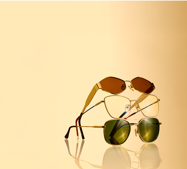 Stack of three gold-framed sunglasses with different lens tints—brown, clear, and green—on a reflective surface against a warm golden background.