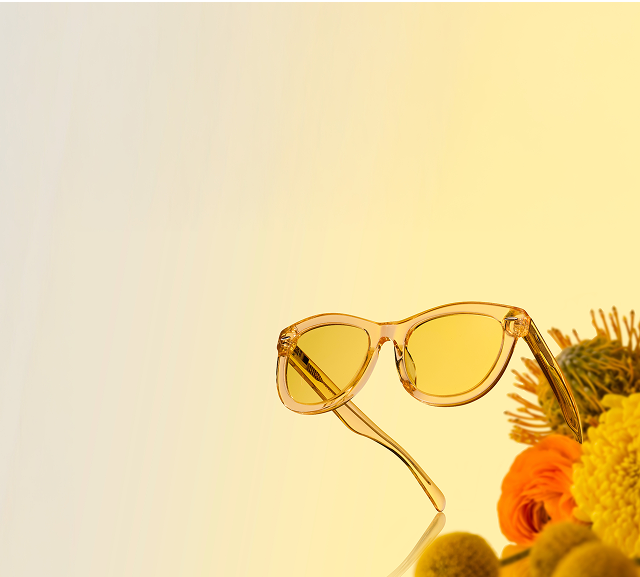 Translucent yellow sunglasses on a reflective surface with yellow and orange flowers in the background.