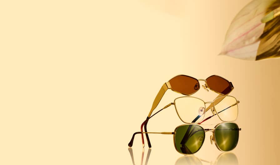 Stack of three gold-framed sunglasses with different lens tints—brown, clear, and green—on a reflective surface against a warm golden background.