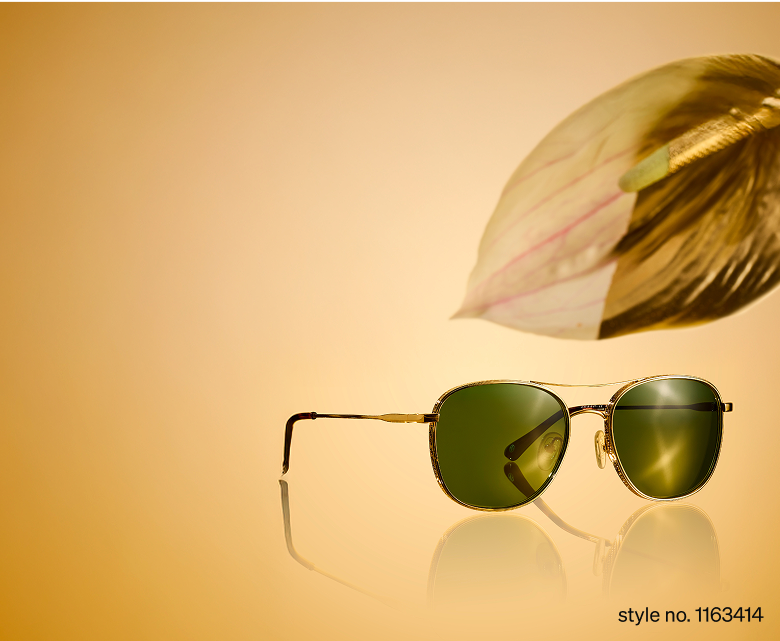 Gold sunglasses style no. 1163414 with dark green lenses on a reflective beige surface next to a blurred leaf.