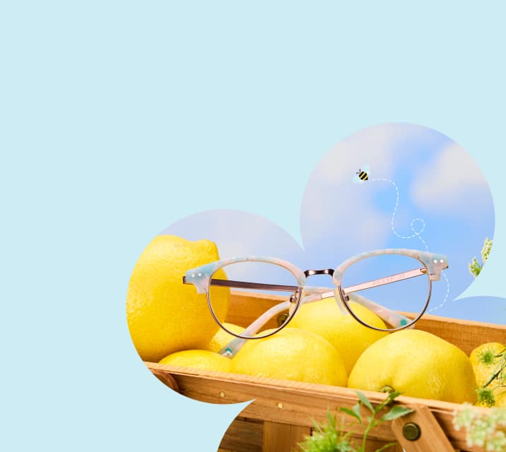 A pair of round, light blue and white marbled browline glasses with rose gold temples resting in a wooden crate filled with bright yellow lemons against a pale blue sky with a small cartoon bee.
