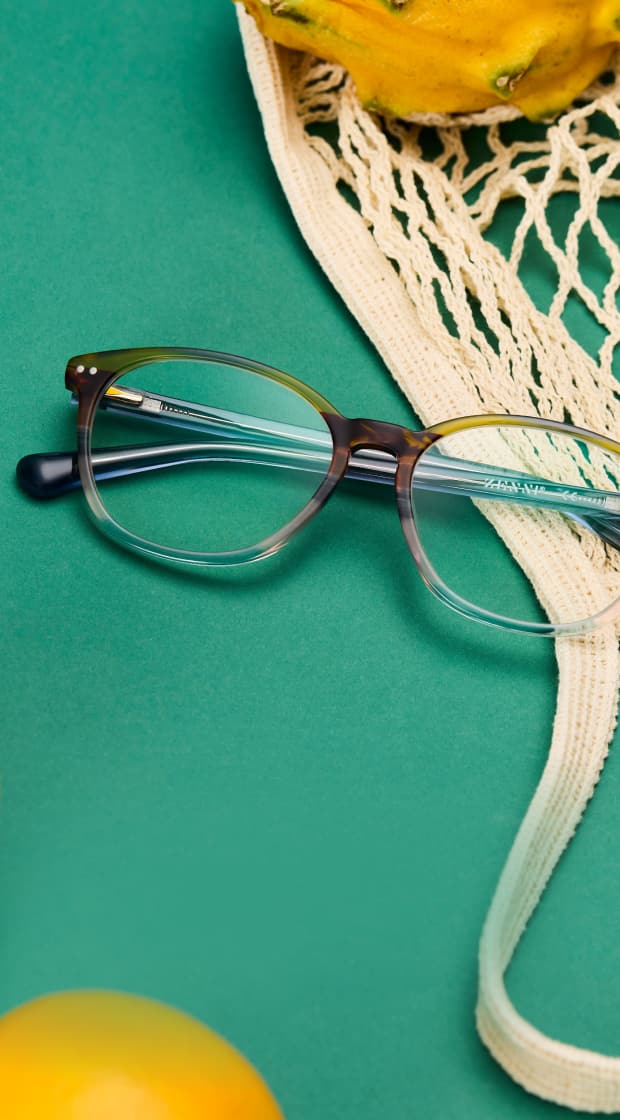 A pair of round glasses with a subtle grey-to-clear gradient resting on a green surface next to lemons and a mesh shopping bag.