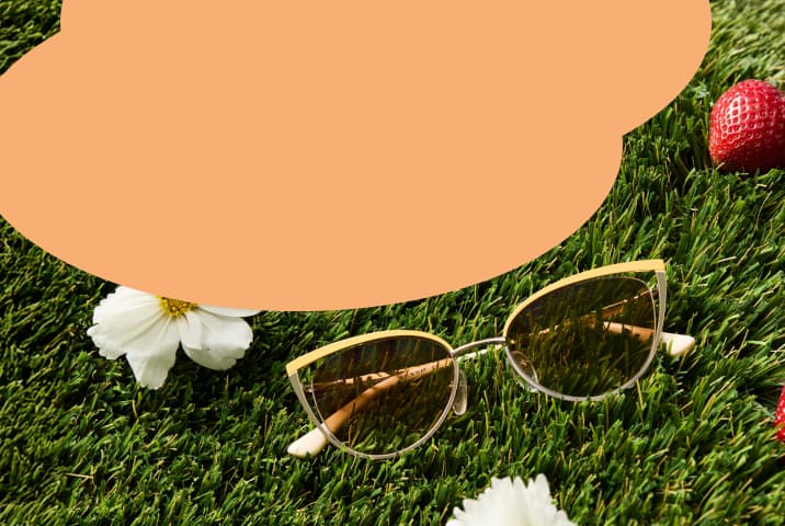 A pair of cat-eye sunglasses with yellow frames and amber-tinted lenses resting on green grass next to white daisies.