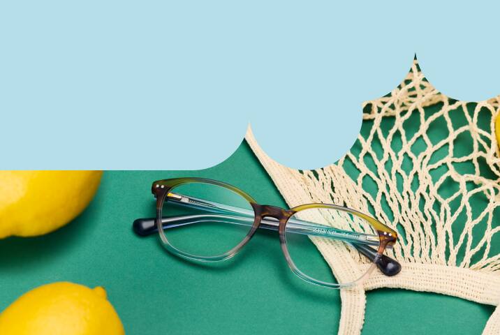 A pair of round glasses with a subtle grey-to-clear gradient resting on a green surface next to lemons and a mesh shopping bag.