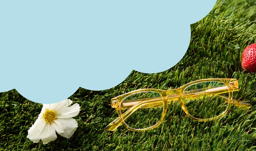 A pair of translucent yellow rectangle glasses resting on lush green grass next to a white daisy and a single red strawberry, with a large light blue cloud-shaped cutout in the upper left corner.