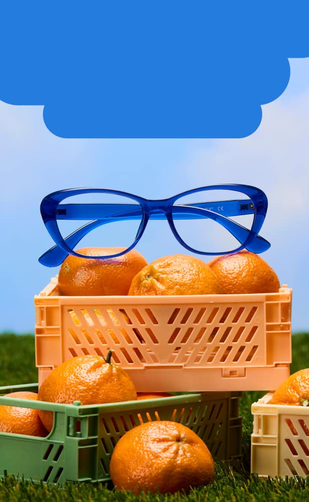 A pair of vibrant blue cat-eye glasses resting on top of a crate of oranges in a field of green grass under a clear blue sky.