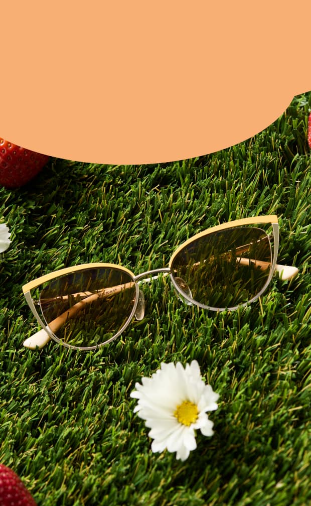 A pair of cat-eye sunglasses with yellow frames and amber-tinted lenses resting on green grass next to white daisies.