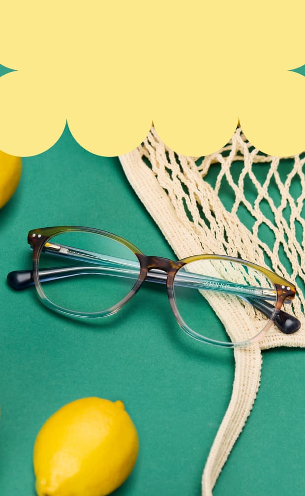 A pair of round glasses with a subtle grey-to-clear gradient resting on a green surface next to lemons and a mesh shopping bag.