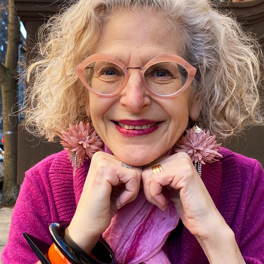 Image of an older woman wearing pink lip-shaped glasses, with a magenta cardigan, light purple scarf, and matching earrings, placing her chin in her hands.