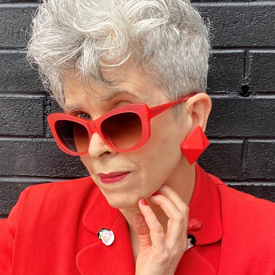 Image of a woman with gray hair wearing an all-red outfit, including her glasses, against a black brick wall.
