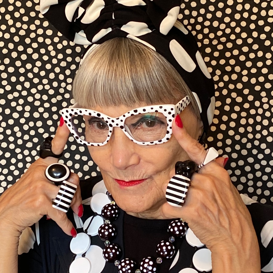 Image of a woman wearing all black and white, wearing polka-dotted glasses in front of a polka-dotted wall.