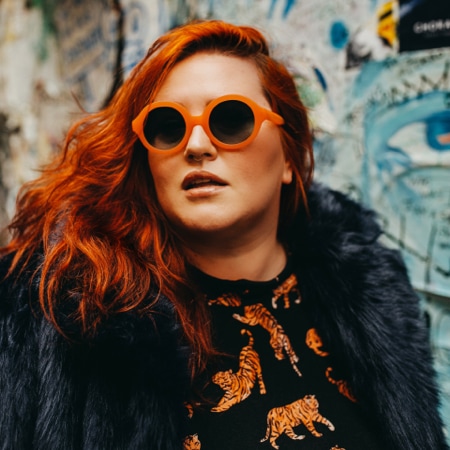 Jessy Parr wearing Zenni Pico round sunglasses #4422225 in front of a piece of colorful artwork.