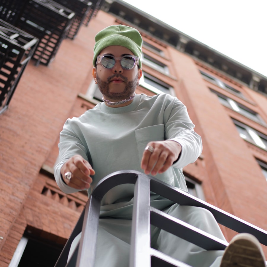 Image of a man wearing a light green outfit and beanie, with a pair of mirrored square sunglasses standing over a handrail.