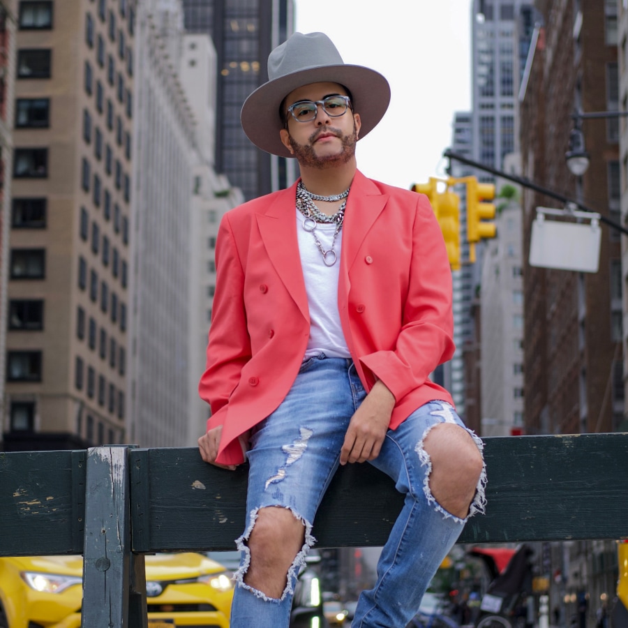 Image of a man wearing a coral-colored coat and ripped jeans, sitting on a handrail. He is wearing a hat and a pair of round patterned glasses.