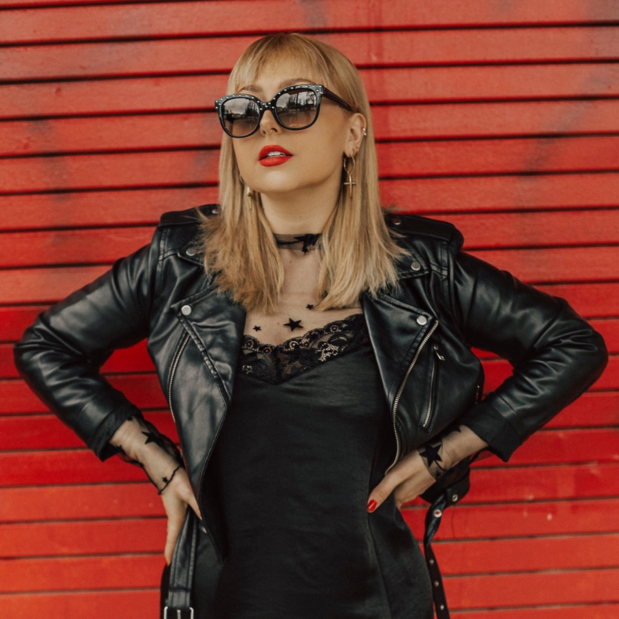 Image of a woman wearing a black leather jacket, with oversized cat-eye sun on. She is standing in front of a red wall.