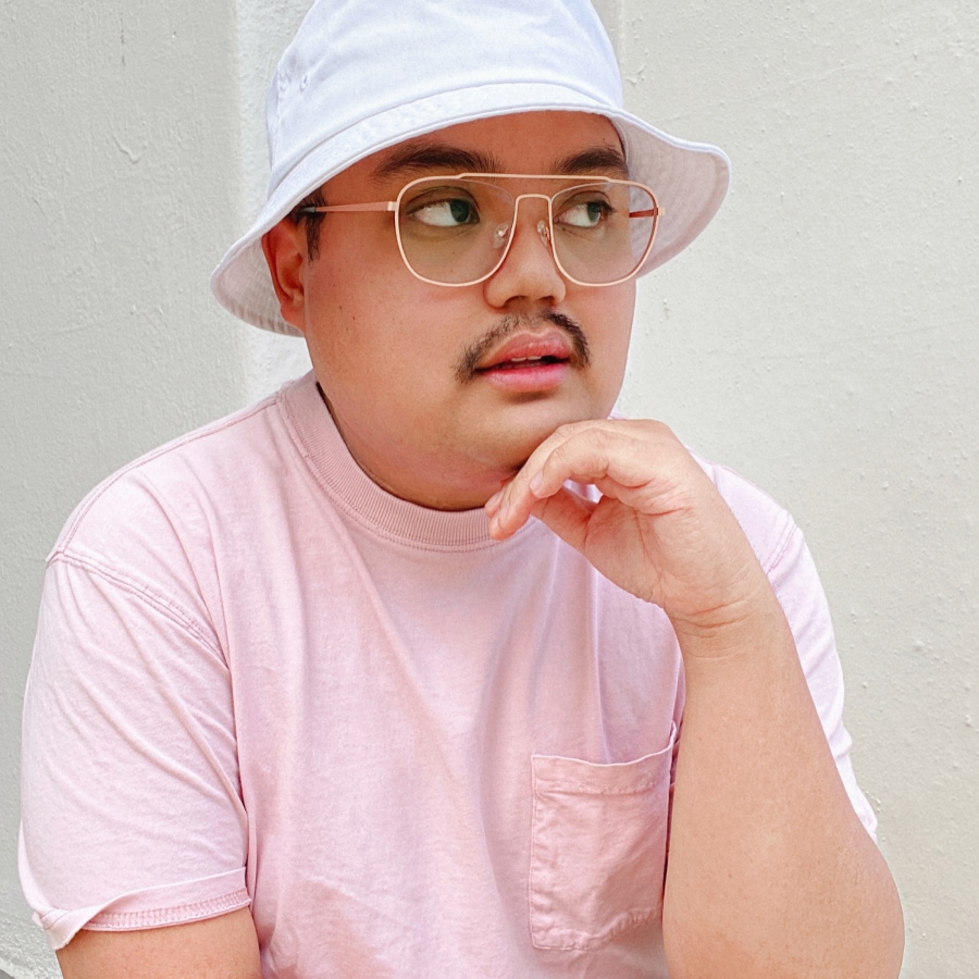 Image of a man wearing a pink tee shirt and matching rose gold aviator glasses, with a white hat on.