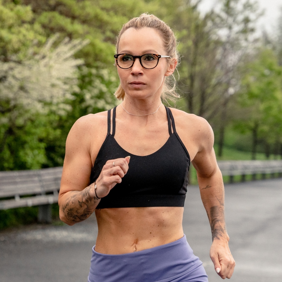 Image of a woman jogging, wearing workout gear and a pair of tortoiseshell round glasses on a runner’s path.