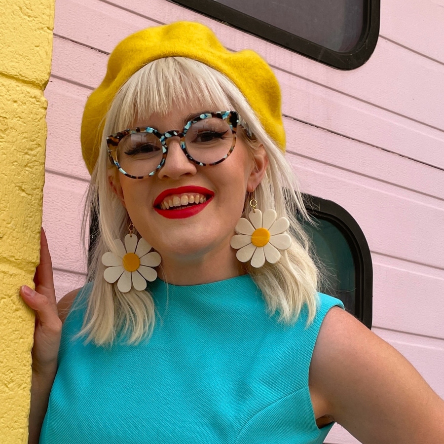 Image of a woman wearing yellow beret and a light blue sleeveless top, along with round blue tortoiseshell glasses, in front of a pink building.