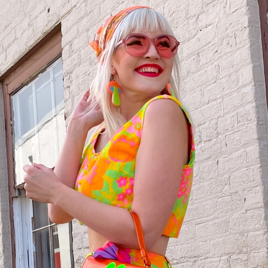 Image of a woman wearing a bright citric color crop top and skirt, wearing pink round sunglasses with pink lenses.