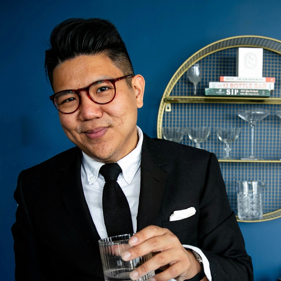 Image of a man wearing a black suit and tie, wearing tortoiseshell glasses holding a cocktail.