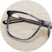Slate blue acetate glasses #4435316 with carbon fiber temple arms folded on top of a light wood tabletop.