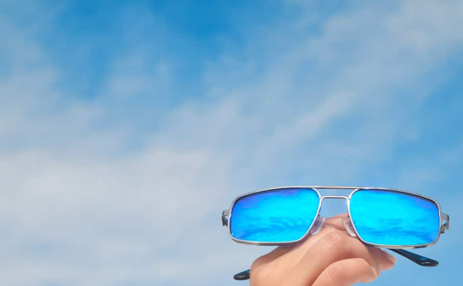 A hand holding silver metal frame aviator sunglasses with blue mirrored lenses.