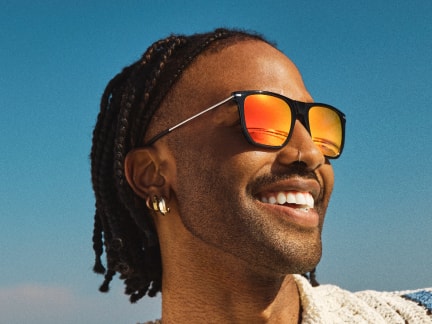 Man with braided hair, small hoop earrings, and a neatly groomed beard, smiling wearing reflective orange sunglasses, looking off to the side against a clear blue sky. The bright sunlight and relaxed expression create a warm, summery vibe.