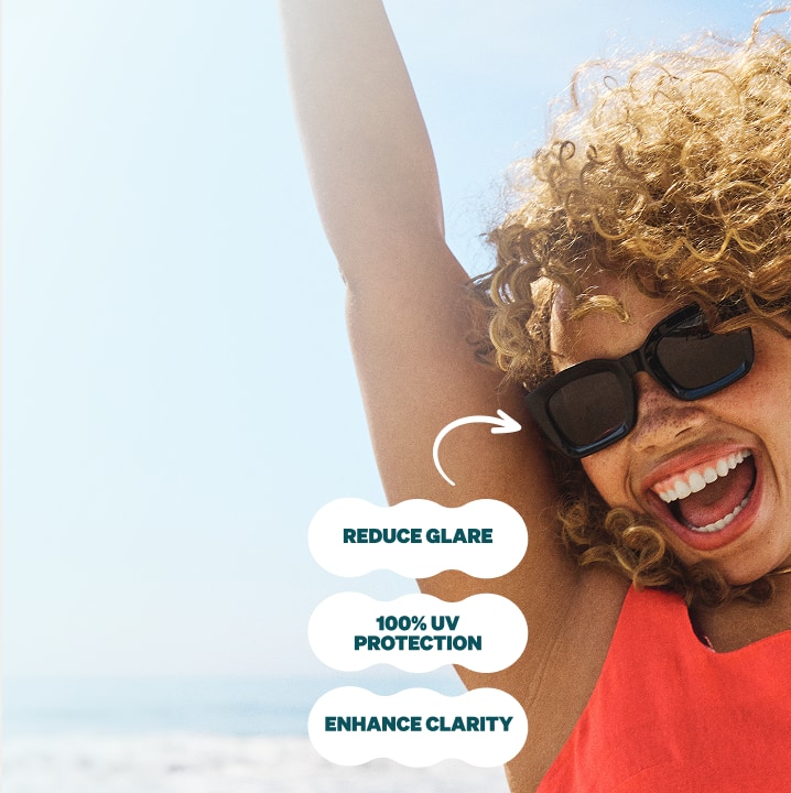 A woman with curly hair at the beach wearing black square sunglasses with gray polarized lenses. Additional text: Enhance clarity, 100% UV protection, reduce glare.