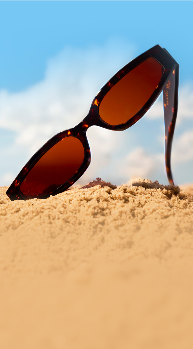 A pair of tortoiseshell sunglasses with brown lenses is partially buried in the sand under a bright blue sky with fluffy clouds.
