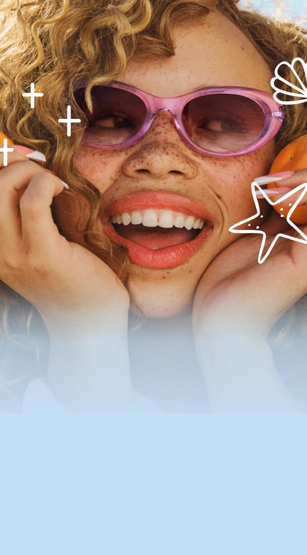 Person with curly hair wearing pink sunglasses smiles with a beach in the background.