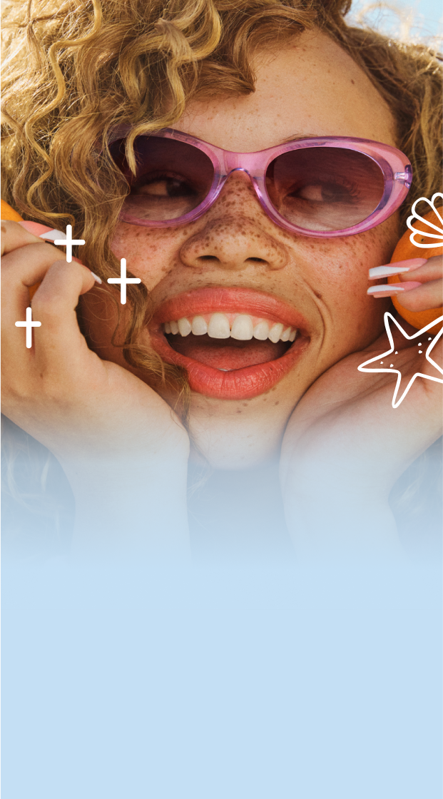 Person with curly hair wearing pink sunglasses smiles with a beach in the background.