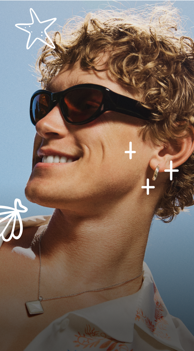 Man with curly, light brown hair, in a light-colored shirt with orange floral patterns, wearing large dark sunglasses, smiling against a backdrop of a cloudless blue sky. 
