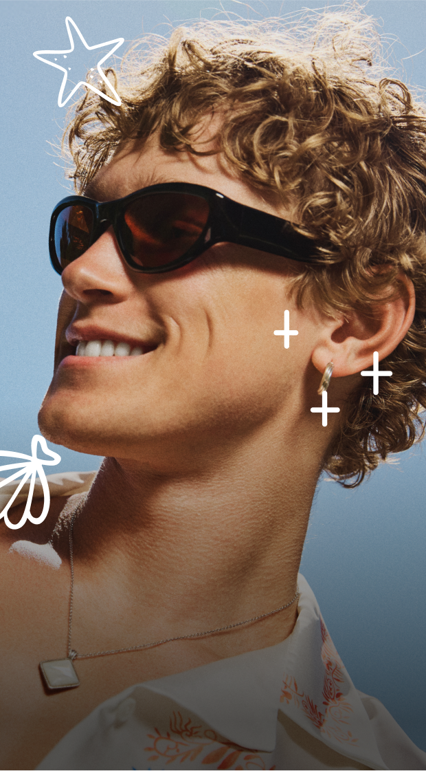 Man with curly, light brown hair, in a light-colored shirt with orange floral patterns, wearing large dark sunglasses, smiling against a backdrop of a cloudless blue sky. 