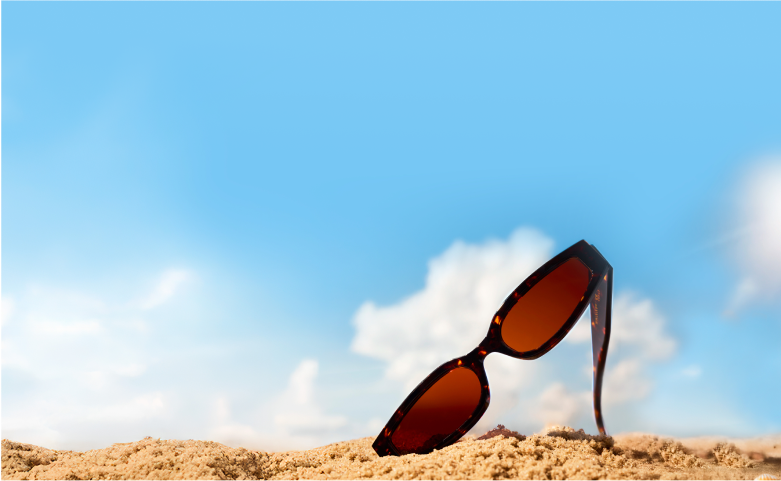 A pair of tortoiseshell sunglasses with brown lenses is partially buried in the sand under a bright blue sky with fluffy clouds.