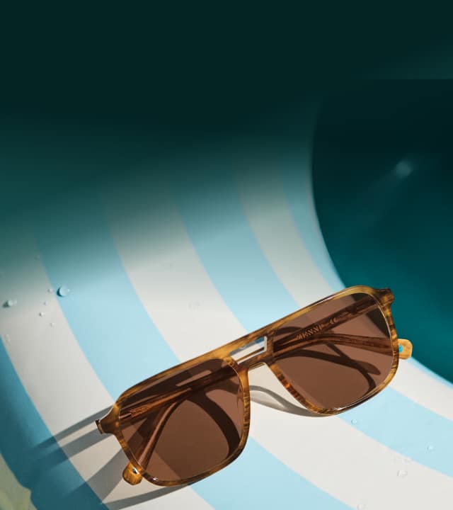 A pair of amber-brown tortoiseshell sunglasses with dark lenses resting on a light blue and white striped pool float, with water droplets adding a refreshing, summery vibe under soft lighting.