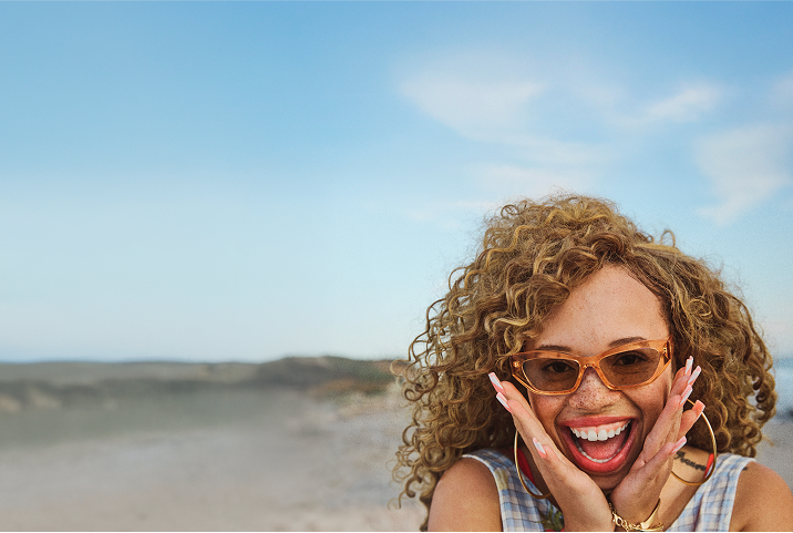 A smiling person with curly hair and brown-tinted sunglasses holds their face with both hands on a sunny beach, with sand, hills, and a blue sky in the background.