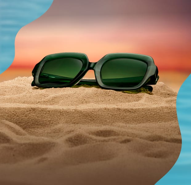 A pair of thick green rectangular sunglasses resting on a mound of sand against a sunset-colored sky and blue water..