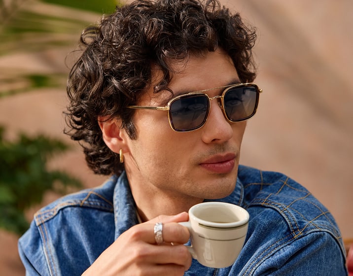 A man in gold square aviator Zenni sunglasses. He has mid-length curly brown hair and is sipping an espresso.